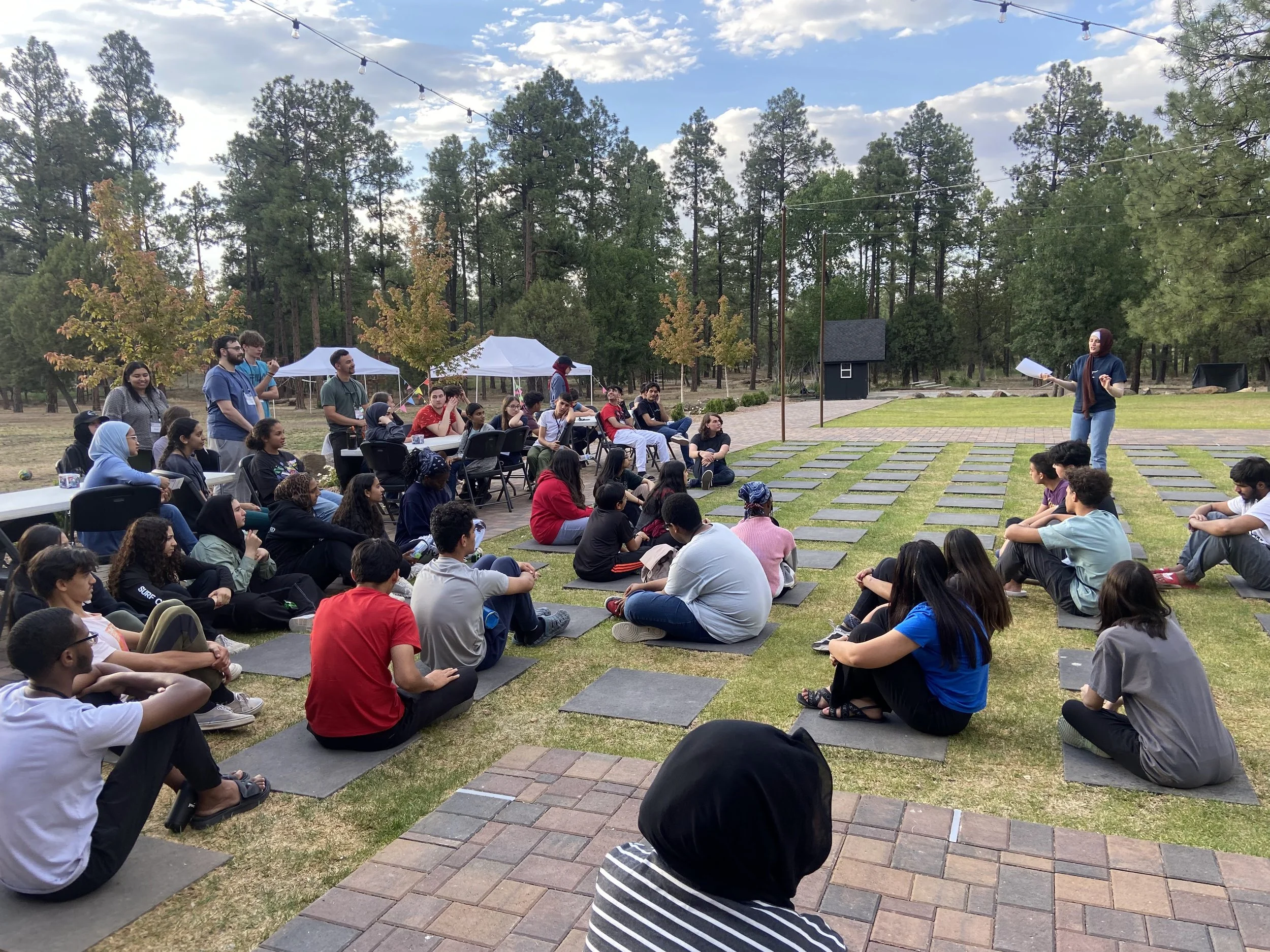 An outdoor gathering of diverse people sitting on mats in front of a woman speaking to a group, with trees, tents, and string lights overhead in a park-like setting.