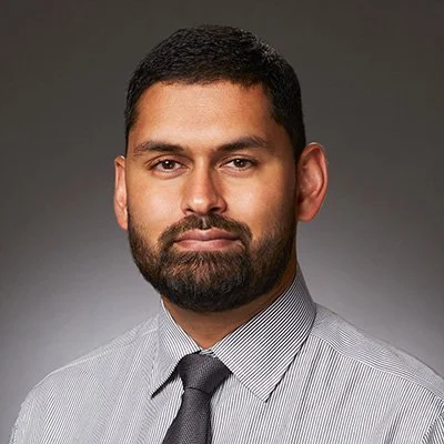 Portrait of a man with dark hair and beard wearing a light gray dress shirt and gray tie against a plain gray background.