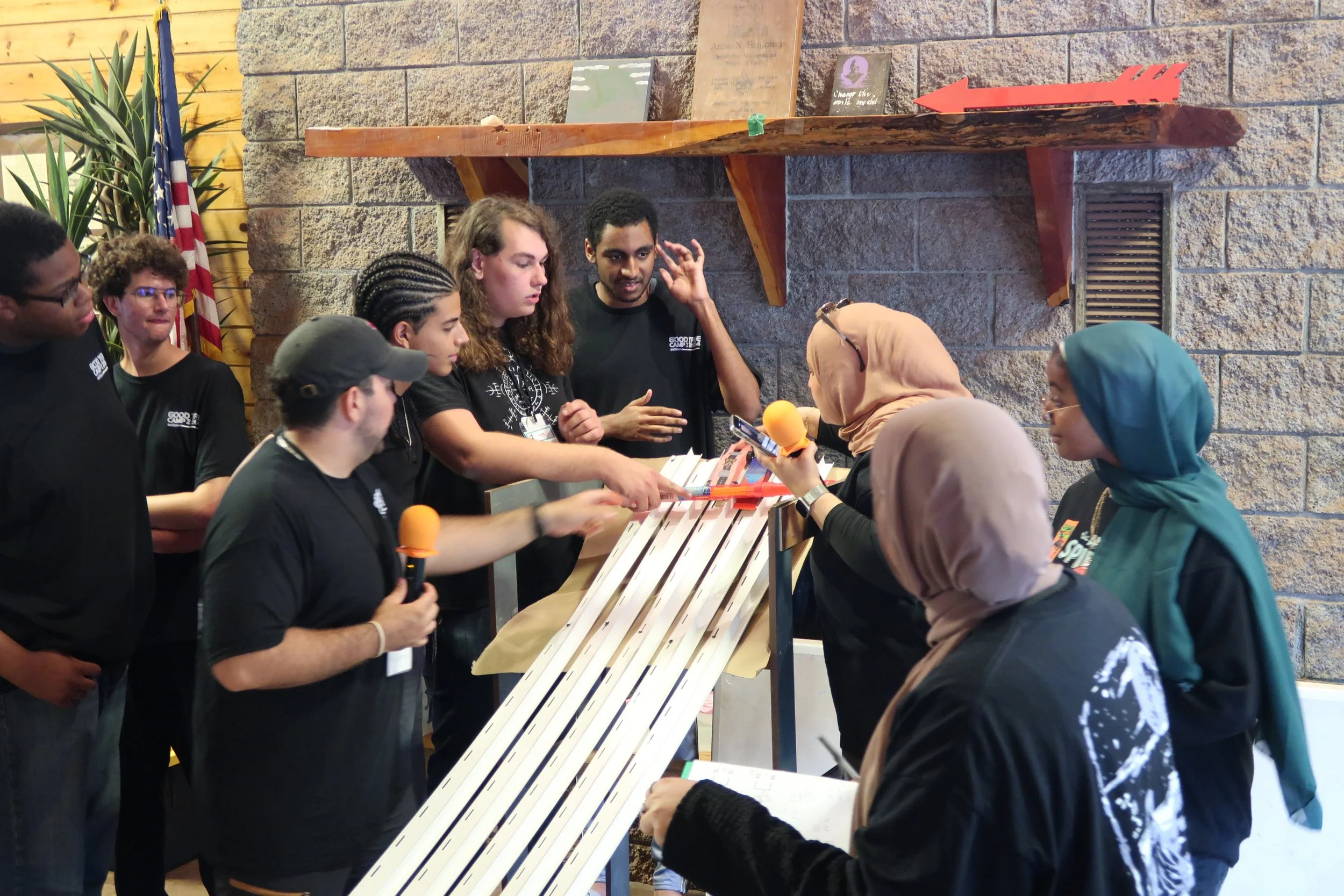 A group of young people, some wearing hijabs, participating in a team activity involving a large wooden ramp with marbles, with one person holding an orange microphone and others observing and discussing.
