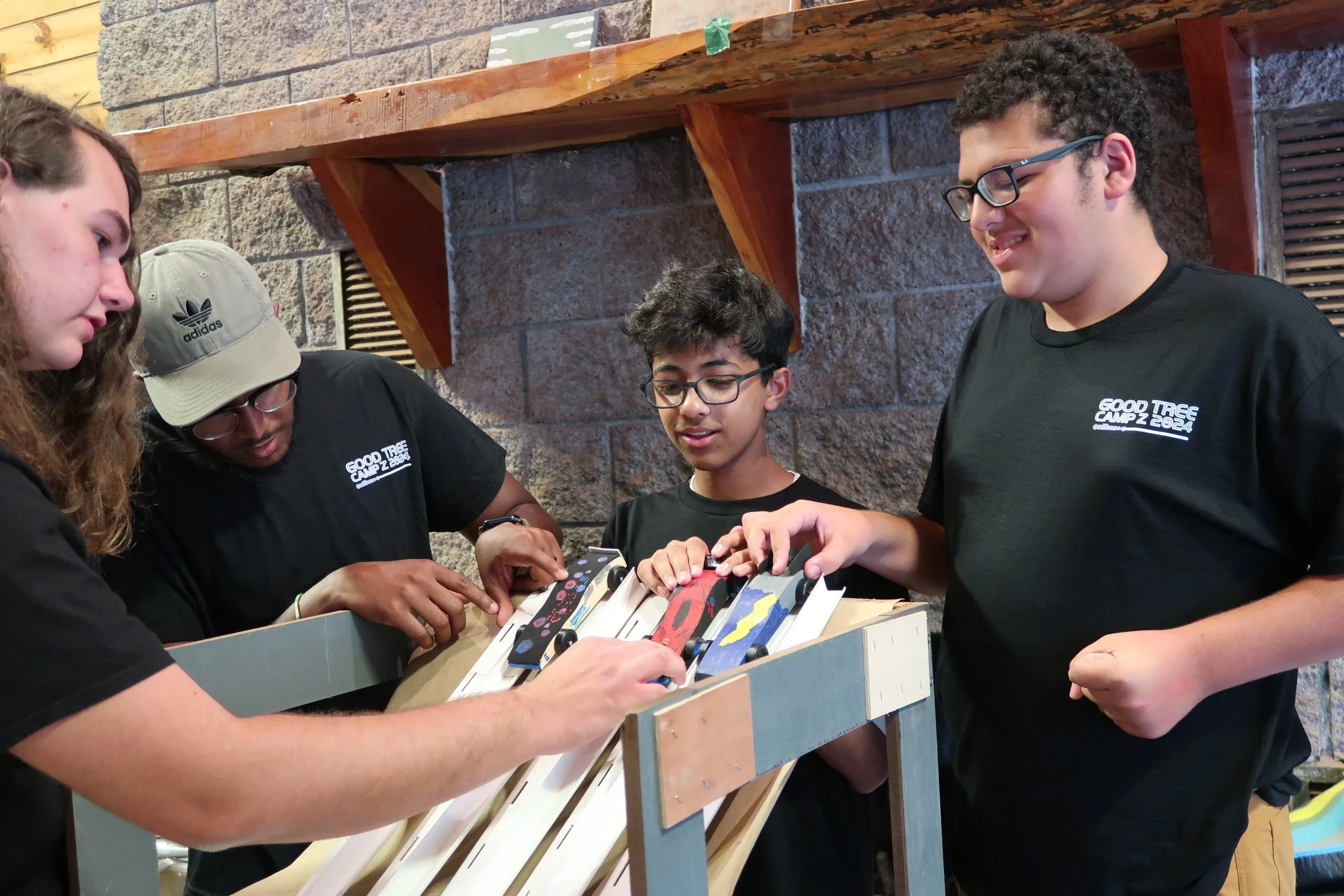 Four teenagers are gathered around a wooden ramp, testing toy cars with different designs, in an indoor setting with brick walls and wooden shelves.