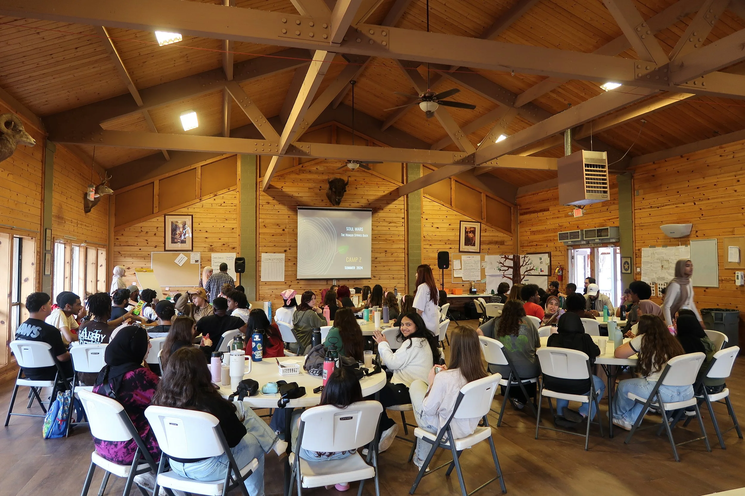 Large group of young people gathered in a wooden banquet hall, sitting at round tables, some facing a projector screen at the front of the room.