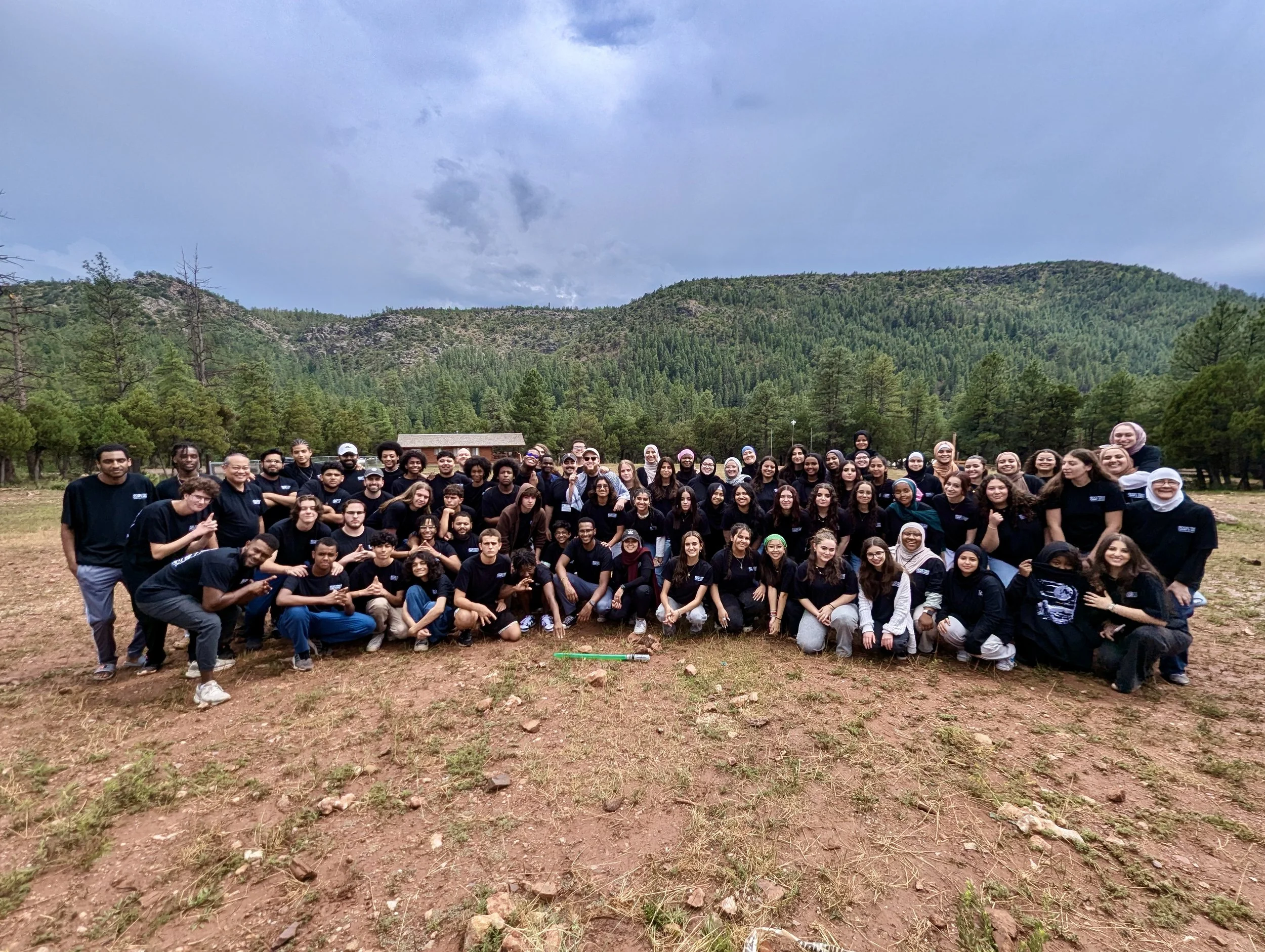 A large group of diverse young people posing outdoors on a dirt field with green trees and mountains in the background.