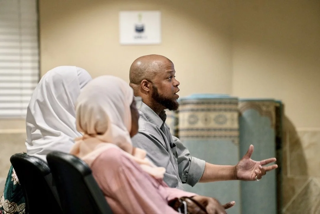 A man speaking while seated in a meeting room, flanked by two women wearing white hijabs, with a blurred background and a window with blinds to the side.