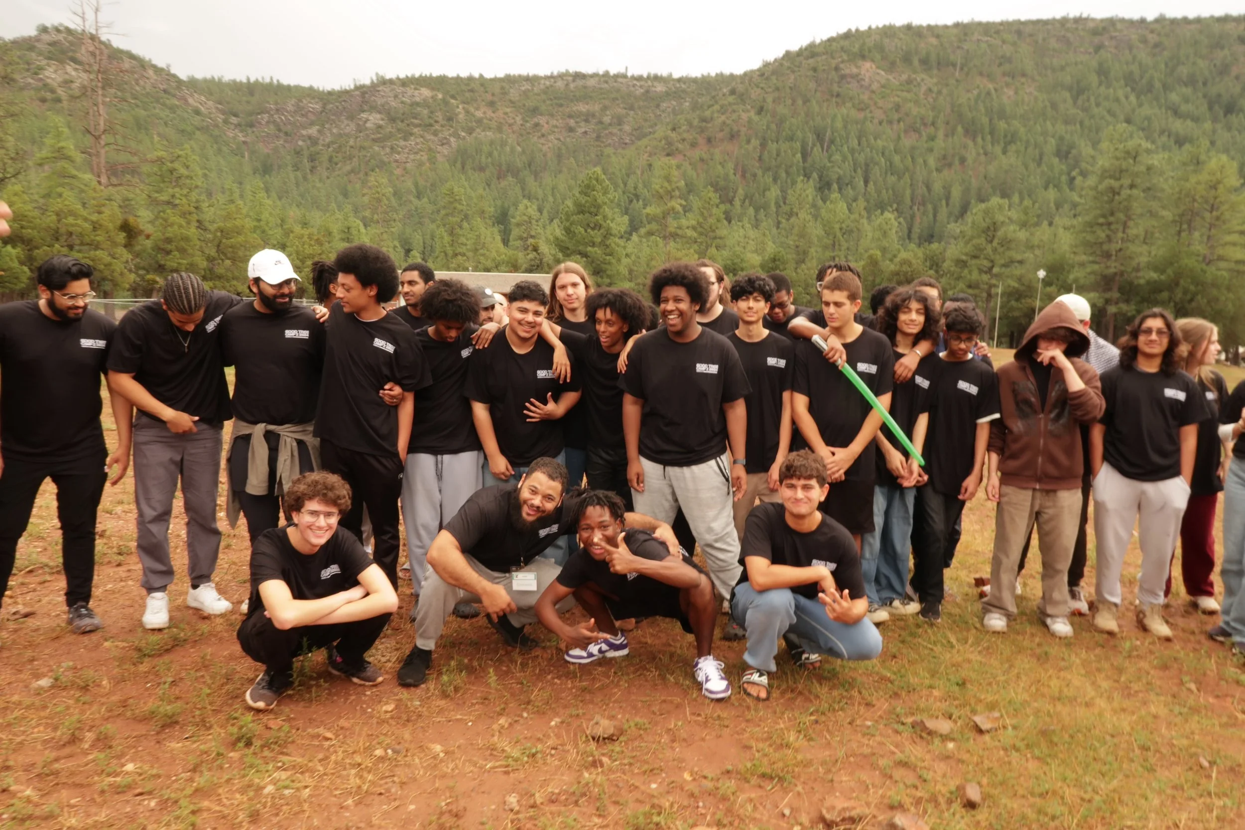 A diverse group of young people gathered outdoors on a grassy area with a forested hillside in the background, some wearing black t-shirts and smiling.