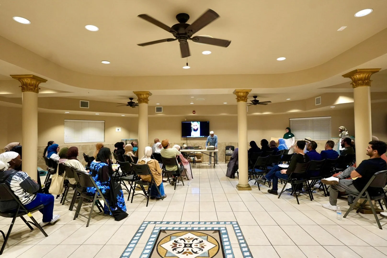 A diverse group of people seated in a semicircular arrangement in a spacious indoor venue, listening to a speaker at a table in front of a large screen displaying an image. The room has beige walls, decorative columns with gold accents, ceiling fans, and tiled flooring with a decorative pattern in the center.
