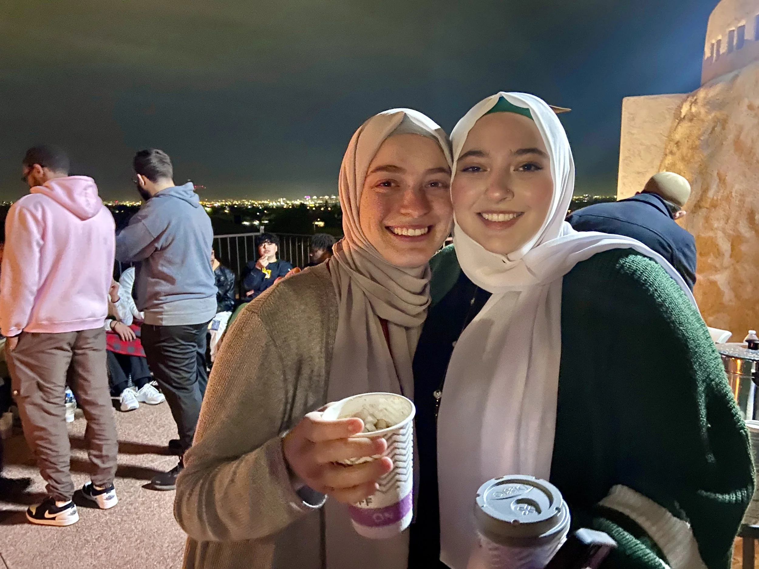Two young women wearing headscarves smiling at night, holding cups of ice cream and coffee on an outdoor terrace with a city skyline in the background, other people gathered nearby.