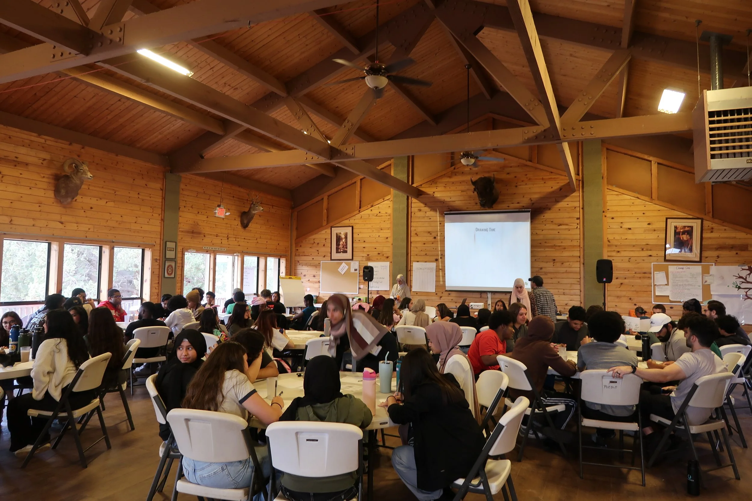A large group of people sitting at round tables in a wooden hall with mounted animal heads on the walls, large windows on one side, and a presentation screen at the front.