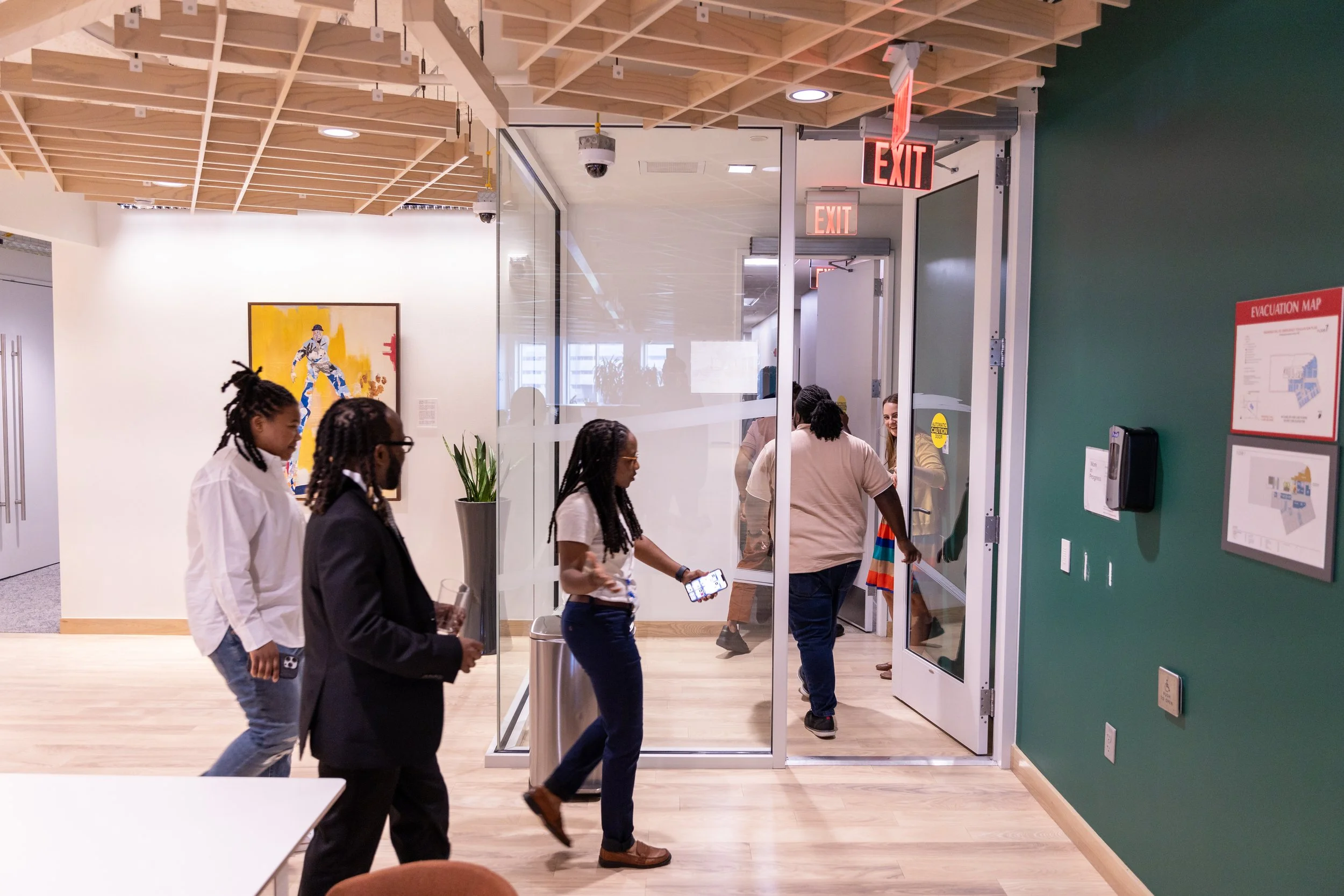 People walking through a modern office hallway with glass doors and an EXIT sign.
