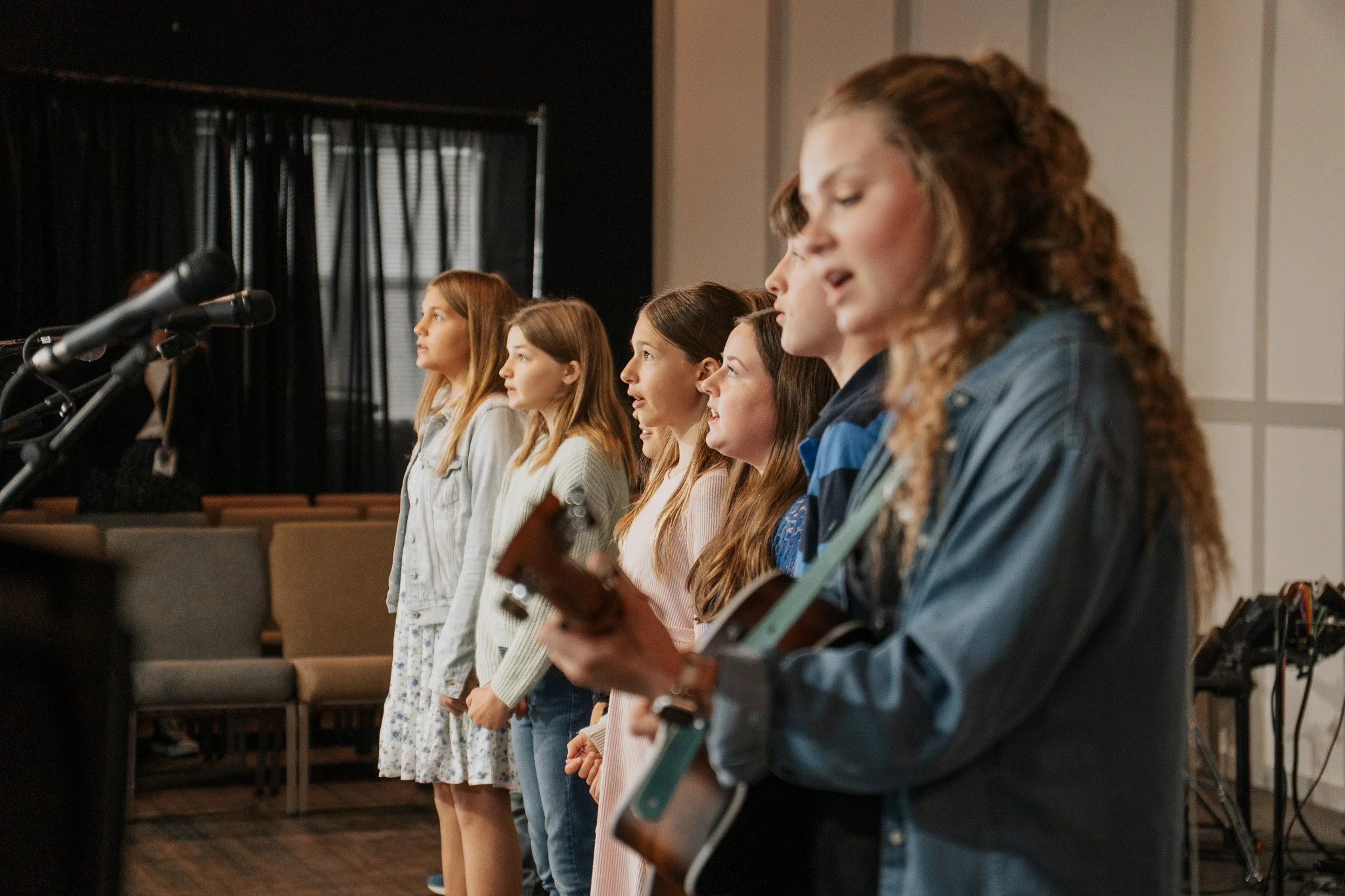 Six young women stand in a line, singing and playing guitars during a rehearsal or performance in a room with wooden flooring and black curtains.