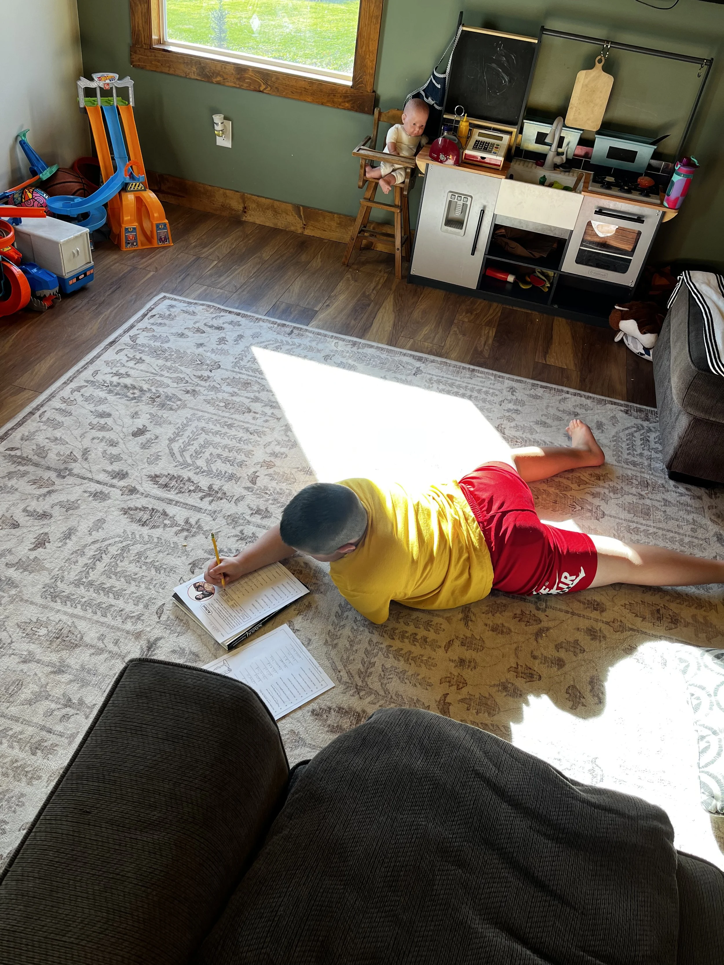 A boy in a yellow shirt and red shorts is lying on a carpet reading a book with a notebook nearby. A younger child is sitting in a wooden high chair near a play kitchen with toy food and utensils.