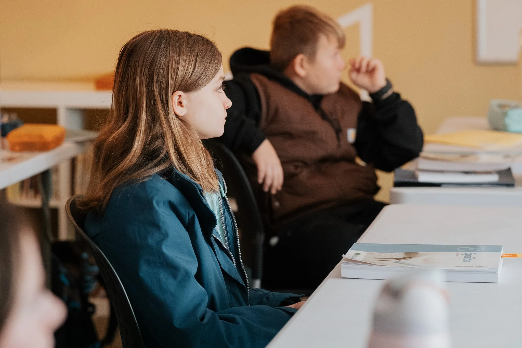 A girl with long, light brown hair sitting at a desk in a classroom, wearing a dark green jacket, with a boy in a black and brown jacket sitting nearby, both looking ahead attentively.
