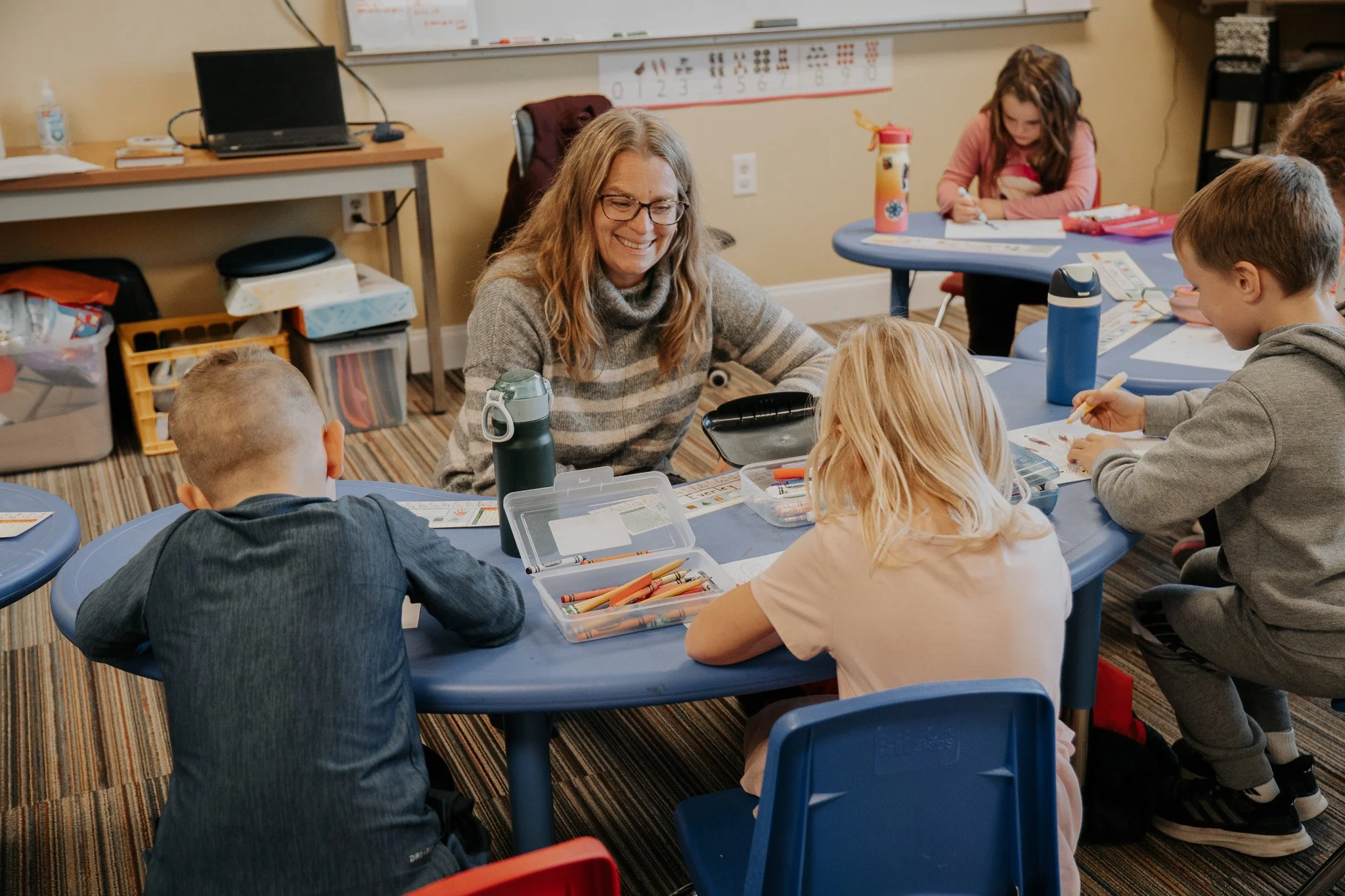 A teacher sitting with a group of elementary school children at a classroom table, smiling and engaging in a learning activity with colored pencils.