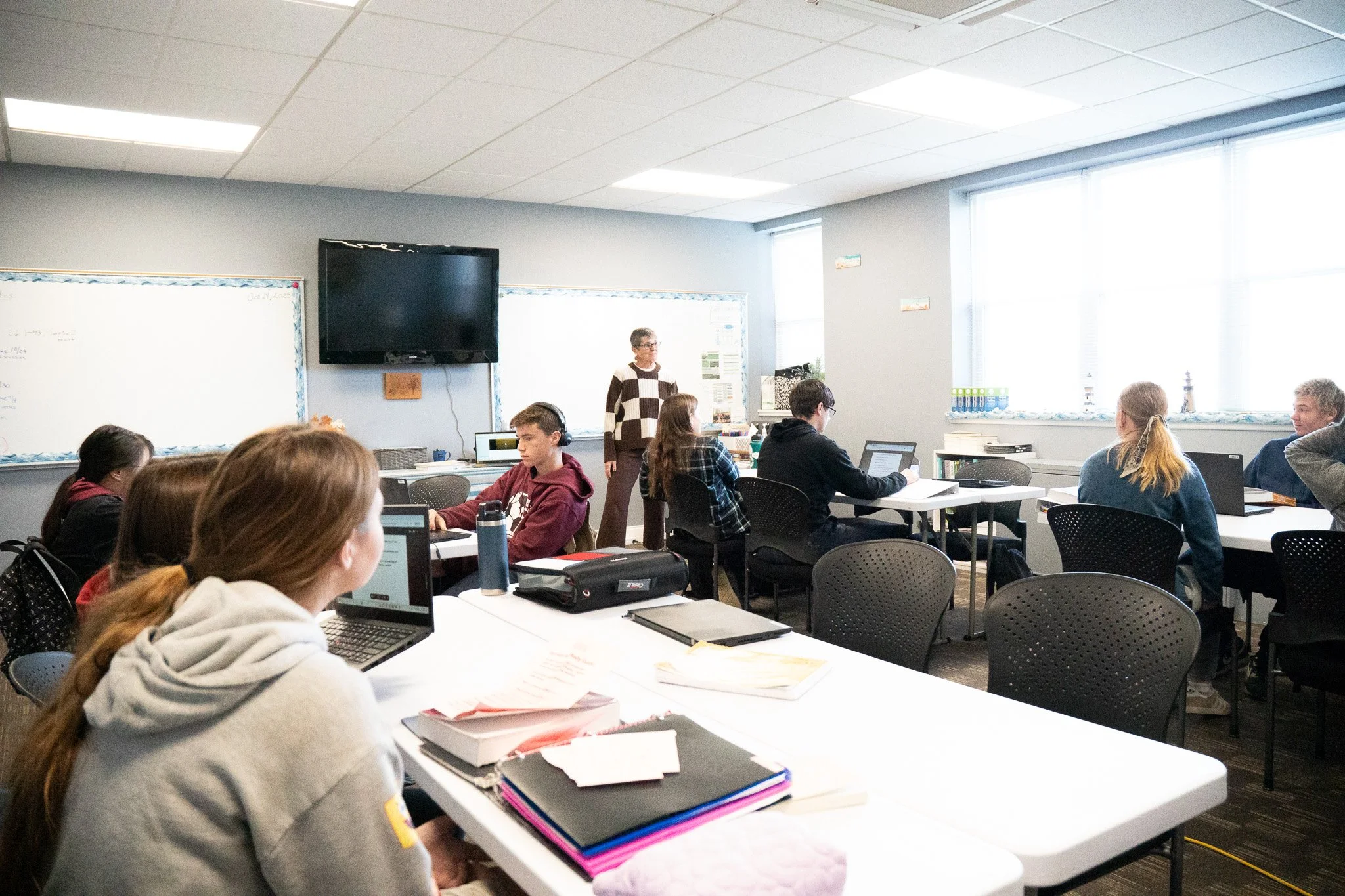 Classroom with students seated at tables using laptops, a teacher standing in front of a whiteboard, and a TV mounted on the wall.