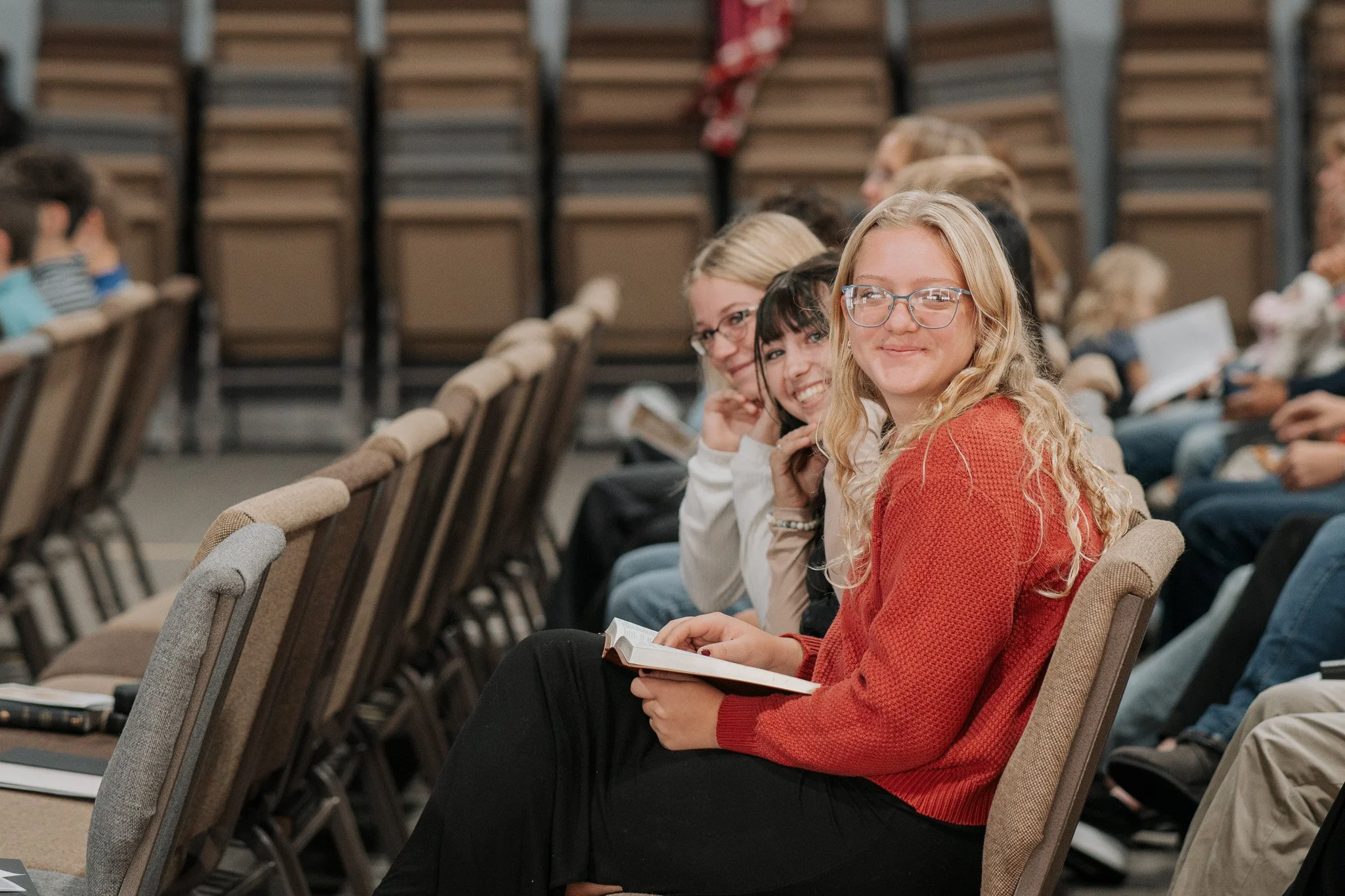 A group of teenagers sitting in an auditorium, smiling and looking at the camera. The girl in the foreground has long, blonde curly hair, glasses, and is holding a book. Others behind her are sitting with happy expressions, some with books or papers.