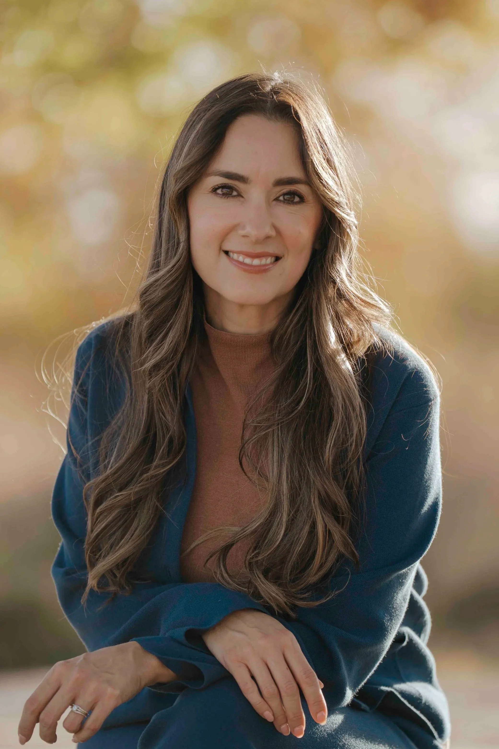 A young woman with long wavy brown hair, smiling, sitting outdoors with blurred autumn foliage in the background.