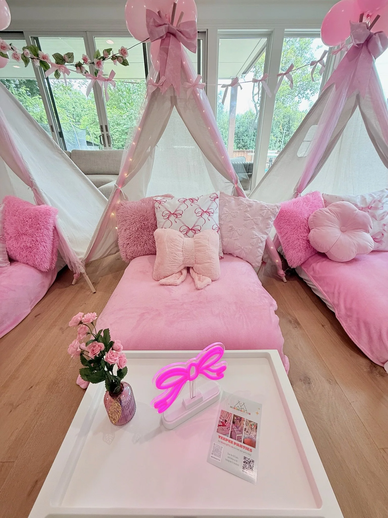 Pink and white themed indoor teepee area with pink pillows, plush cushions, and decorative pillows, featuring a neon pink bow sign and a small vase of pink flowers on a white table, with pastel pink garlands and fairy lights.