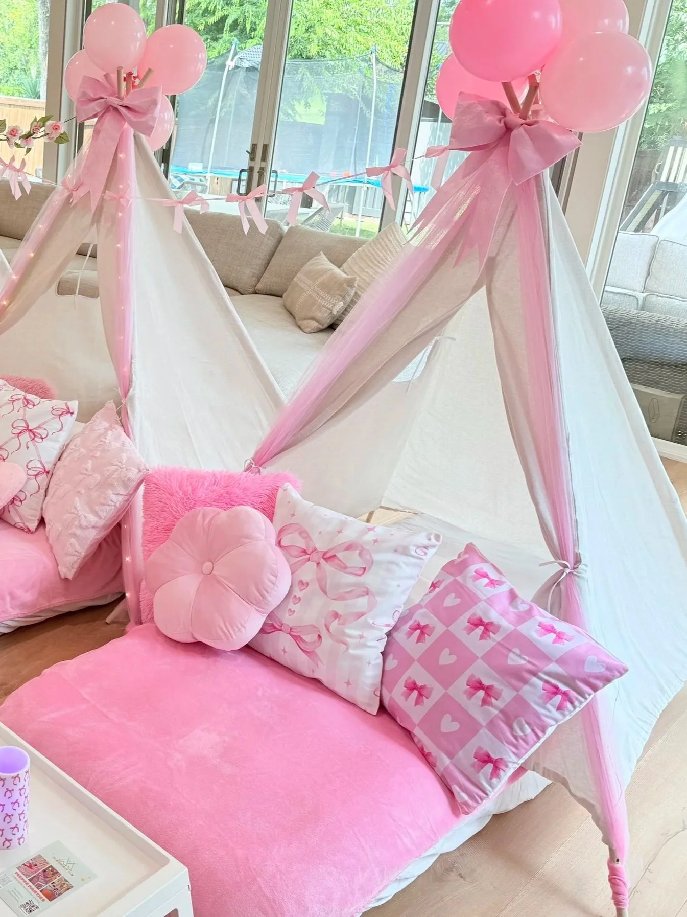 A pink-themed children's bed with a canopy tent decorated with pink balloons and ribbons, surrounded by pink pillows and bedding, in a sunlit room with large windows.