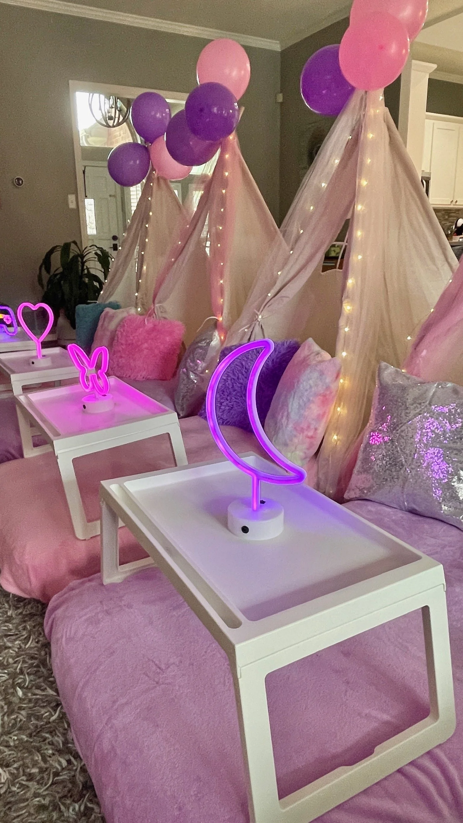 Decorative girl-themed setup featuring pink and purple balloons, white canopy tents with string lights, plush pillows in pastel colors, pink bedspread, and neon moon, butterfly, and heart lights.