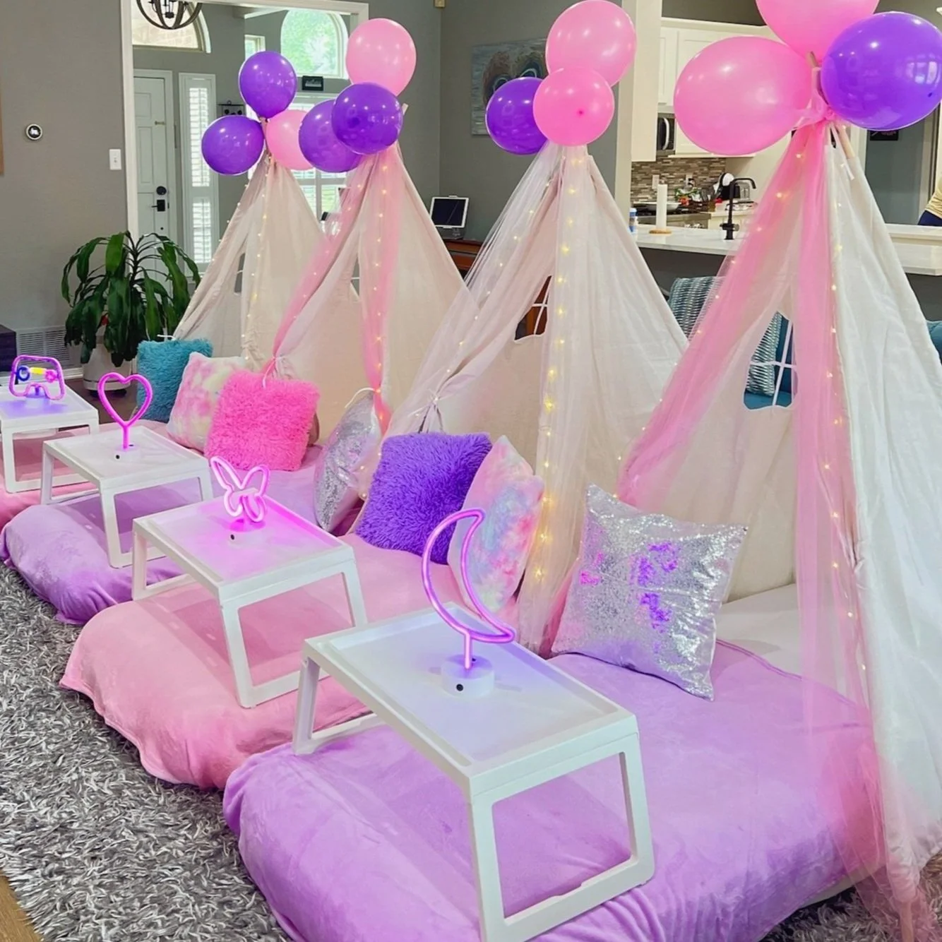 Pink beds with colorful pillows, white tents with fairy lights, purple and pink balloons, neon moon and butterfly lights, on a gray shag rug in a cozy room.