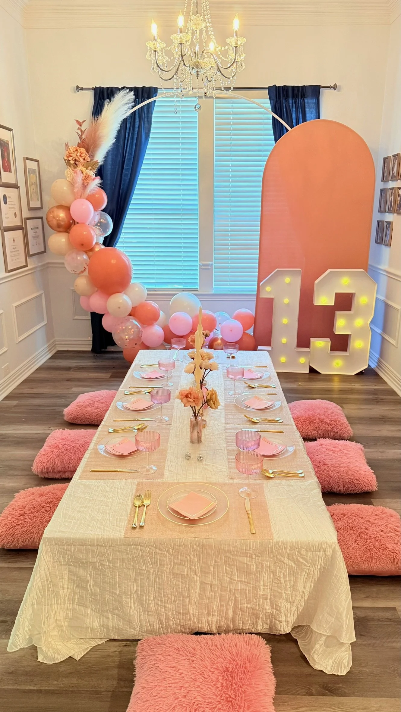 Decorated dining room for a birthday celebration with a long table set with pink napkins, gold utensils, pink glasses, and a centerpiece with pink and white flowers. Behind the table, there's a balloon arrangement in shades of pink, peach, and white, a large pink backdrop, and illuminated numbers 1 and 3 indicating a 13th birthday. A chandelier hangs overhead, and the room has dark blue curtains and framed pictures on the walls.
