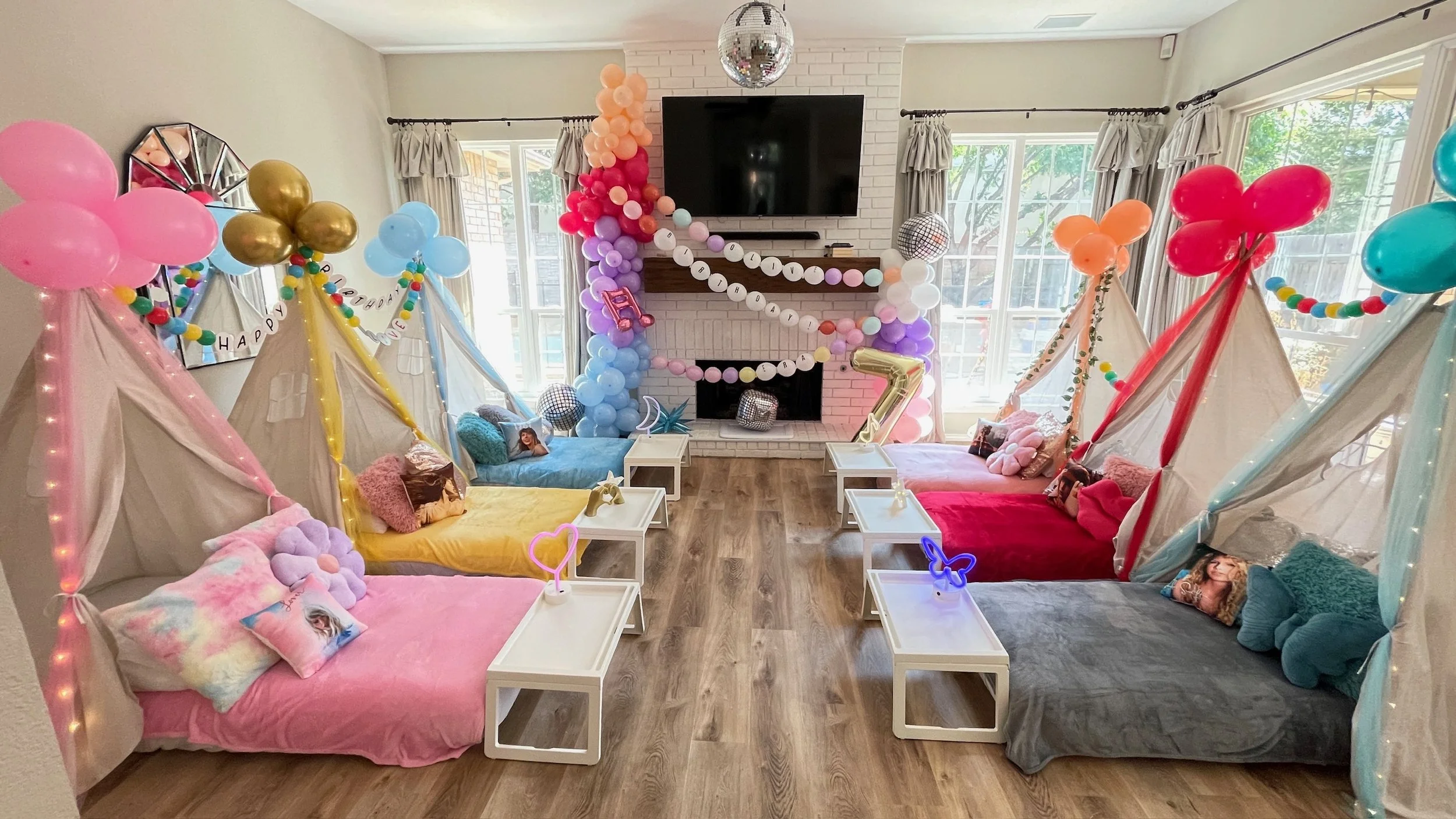 A decorated living room with colorful tents and balloons for a children's birthday party, featuring a yellow, pink, blue, red, and gray tent, with signs and balloon arrangements, a wall-mounted TV, and large windows letting in natural light.