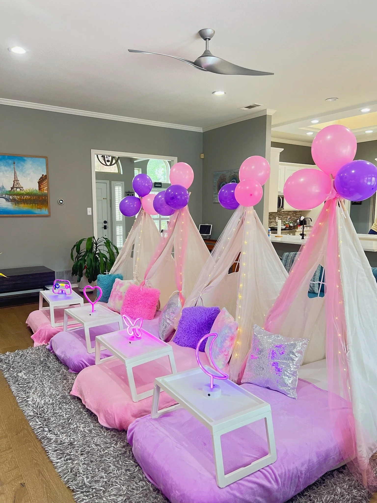 Decorative indoor space with four pink beds, each with pillows, and a canopy of pink and purple balloons and curtains. Neon lights with icons of moon, butterfly, heart, and game controllers are on small white tables in front of each bed.