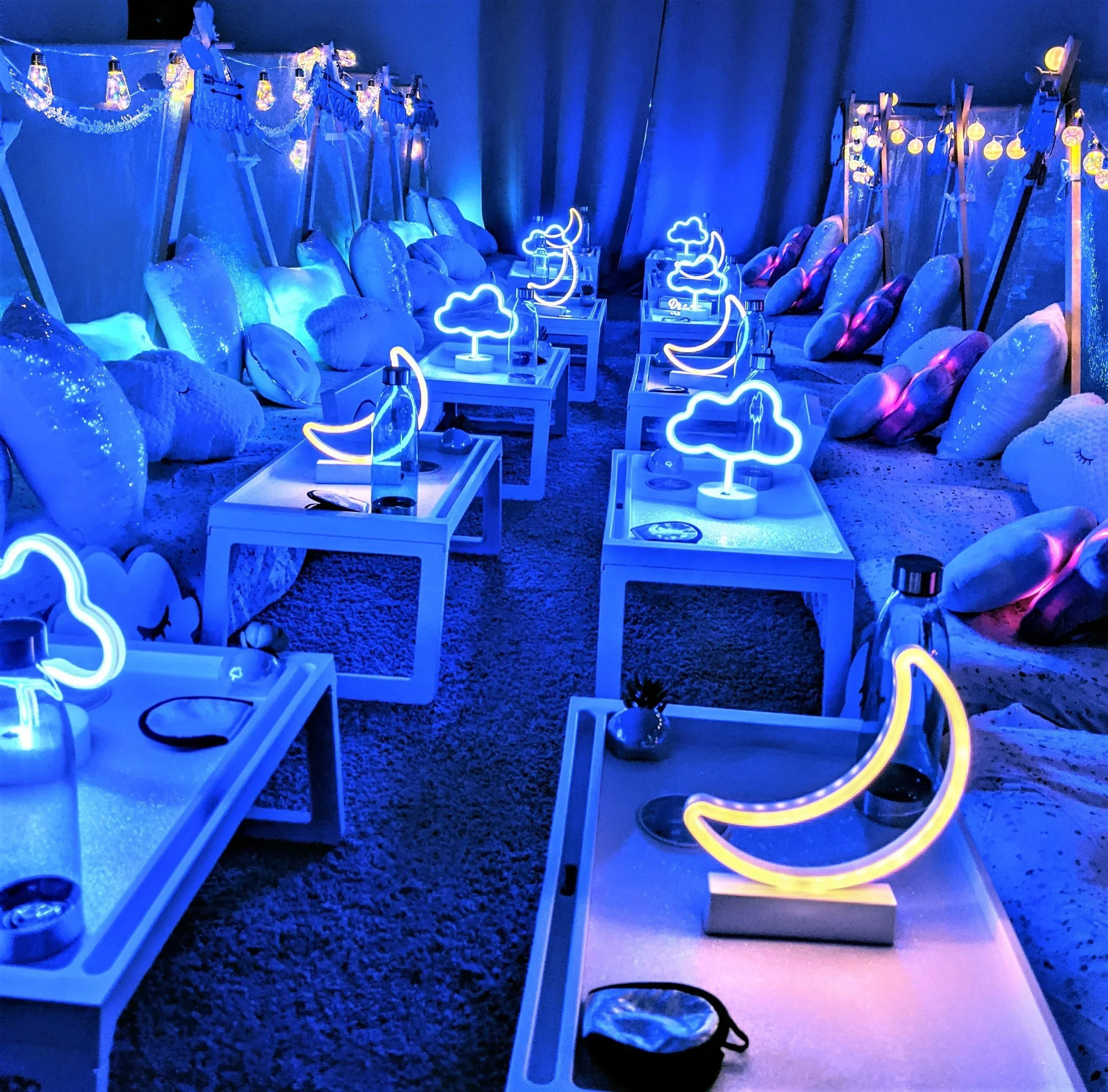 A cozy indoor lounge area with blue neon cloud and moon-shaped lights on white tables, surrounded by plush pillows and stuffed animal clouds on the floor, with blue curtains and string lights overhead.