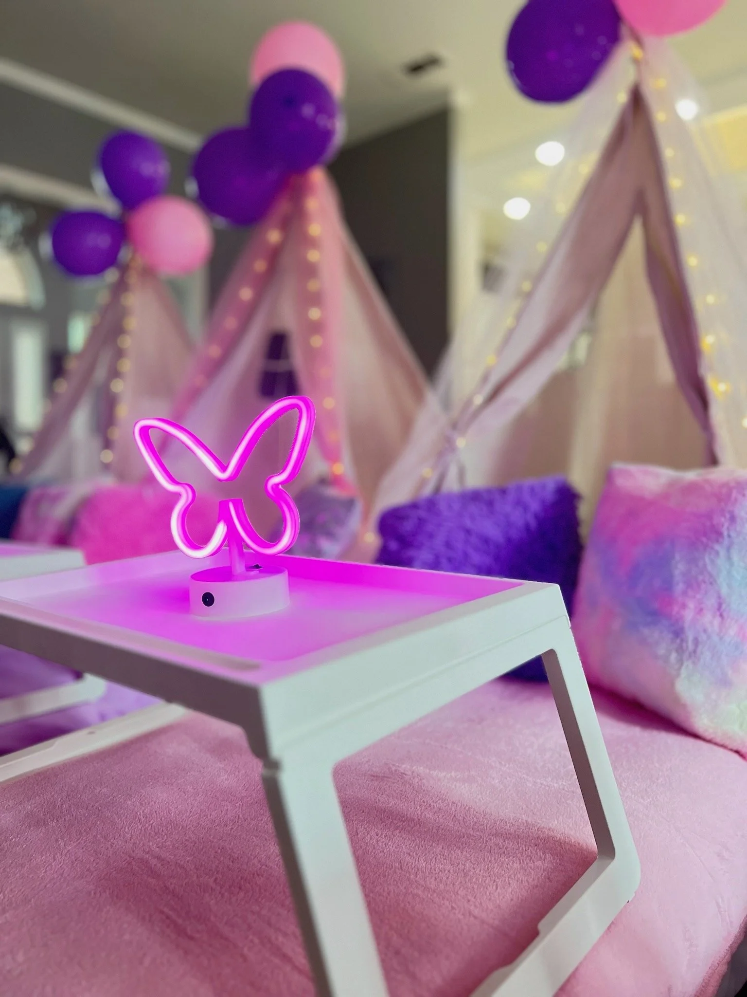 Decorated indoor space with pink and purple balloons, teepees with string lights, colorful pillows, and a pink butterfly-shaped neon light on a white table.