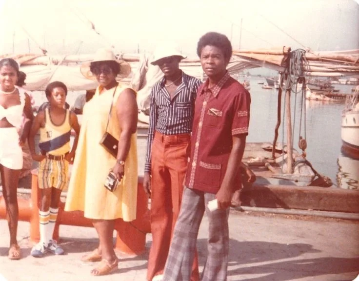 Caribbean family standing by the harbor wearing 1970s leisurewear, including wide-brim hats, patterned shirts, flared trousers, and bright summer dresses.