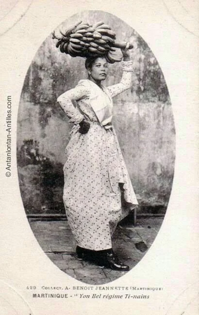 Martinican market woman balancing a bundle of bananas on her head while wearing a traditional Creole dress and headwrap.