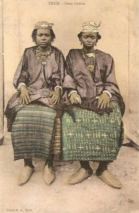 Senegal, circa 1900. Studio portrait postcard of two young Wolof women seated wearing patterned boubou gowns with draped wrapper cloths and headwraps.
