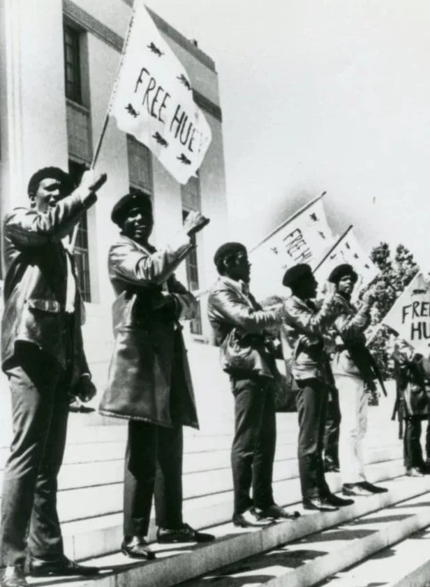 Black Panther members waving Free Huey banners [design by Dolores Davis] on courthouse steps, circa 1960s, Oakland Post Photograph collection, MS 169, African American Museum & Library at Oakland, Oakland Public Library.