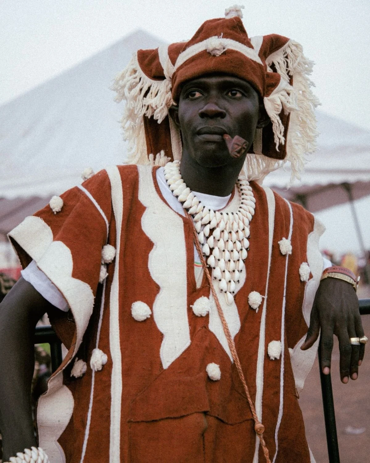 A Dogon man wearing a red and white patterned garment, a tasseled hat, and a layered shell necklace.