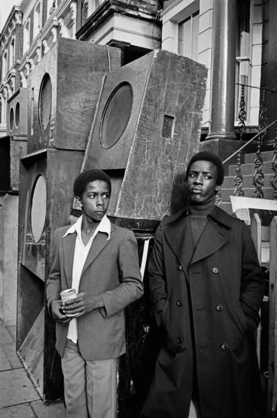 Two young Black men stand beside a stack of large speakers on a residential street,  Notting Hill, London, England, circa 1970s.