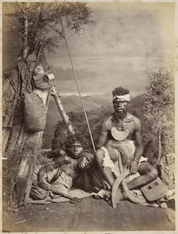 Photo showing a man seated with breastplate and boomerangs, with two women by a bark shelter.