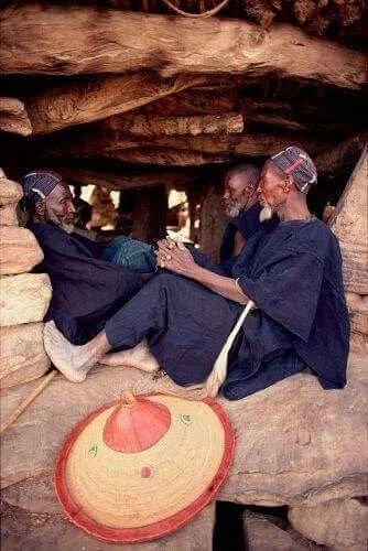 Three Dogon men sit together beneath a rocky overhang in Mali, wearing dark indigo garments and woven straw hats.