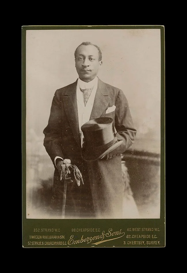 Portrait of a Black man holding a top hat and gloves, posed with a walking stick, conveying middle-class respectability. Photographed by Emberton & Sons Location: London and Surrey, England , circa 1890s.