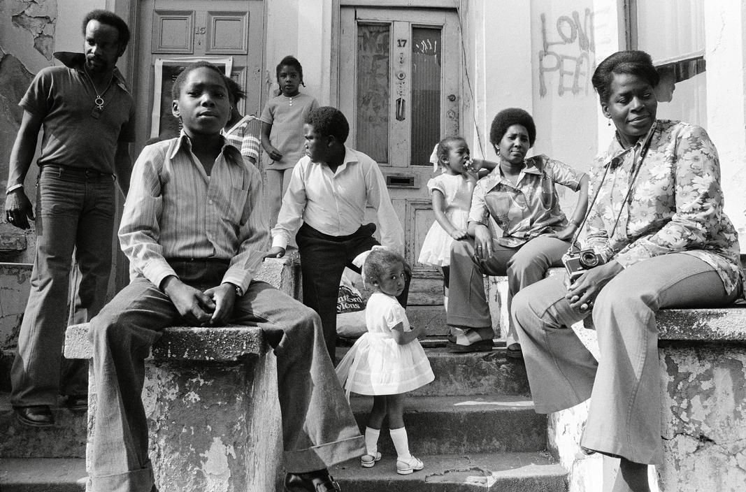 A Black family occupies the steps of a row house. Photographer unknown, West London, England, circa 1970s.