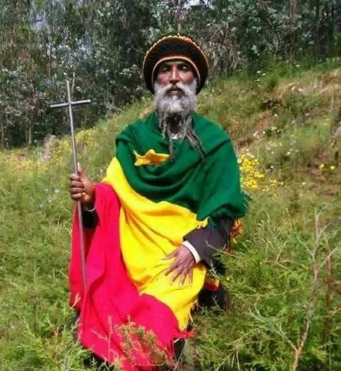 Rastafarian elder draped in the red, gold, and green colors of Ethiopia, holding a cross and wearing a traditional knitted crown associated with Rastafarian identity.