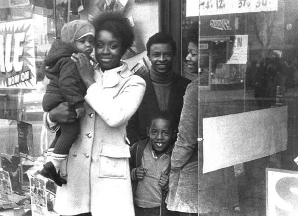 A young Black mother holds a child at her hip while others gather close beside a shop window. Photographed by Charlie Phillips, Notting Hill, London, England, circa 1960s–1970s.