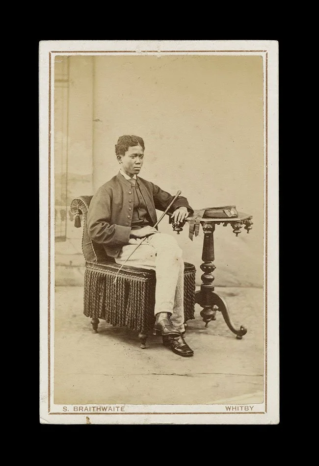 Seated portrait of a young Black boy holding a cane beside a small table, composed in a restrained studio interior. Photographed by S. Braithwaite, Whitby, England , circa 1870s–1880s.
