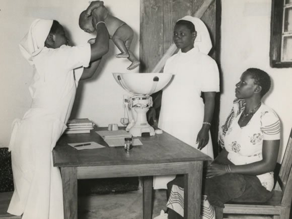 Village midwife, Rachel Yusuti (holding the baby), Mnyiramba and Ayah Brina Masasi, Msukuma, Weighing an infant and giving advice to the mother during an infant welfare clinic session at Kishapu Native Authority Dispensary and Clinic in Shinyanga Dis