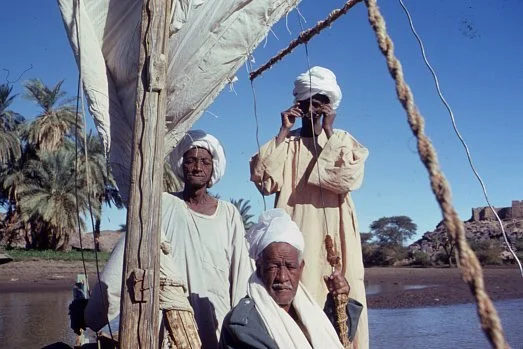 "Nubians in boat - Kulubnarti." Nubian men aboard a traditional river vessel along a palm-lined shoreline. Emery, J. (Photographer). (Untitled photograph). Flickr. © James Emery. Licensed under the Creative Commons Attribution 2.0 Generic (CC BY 2.0)