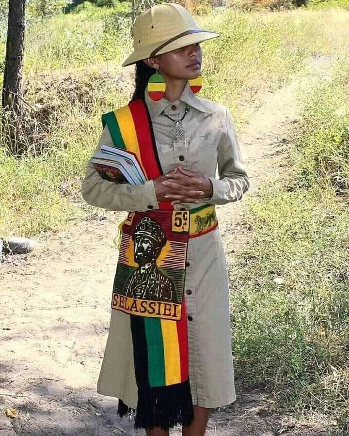 Rastafarian woman wearing a khaki safari dress with red, yellow, and green Selassie I sash and earrings, colors associated with Ethiopian and Rastafarian symbolism.