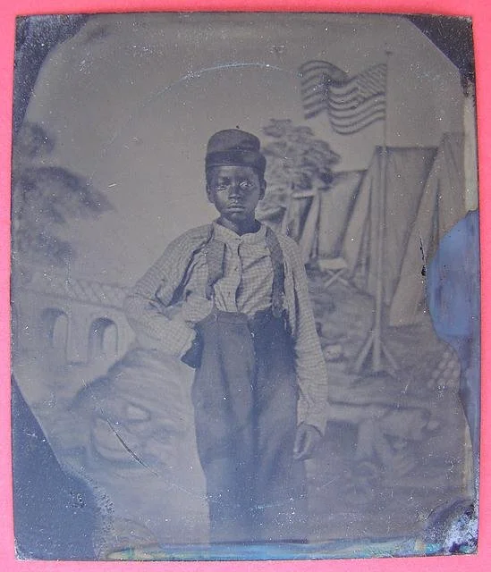 Unidentified African American boy standing in front of painted backdrop showing American flag and tents ; campaign button with portraits of Lincoln on one side and Johnson on the opposite side are attached to inside cover of case.Library of Congress 