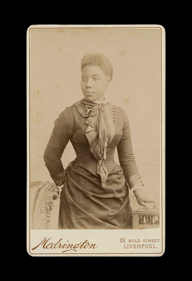 Studio portrait of a Black woman in a fitted dress, posed beside an upholstered chair. Photographed by  Medrington , Liverpool, England , circa 1880s–1890s.