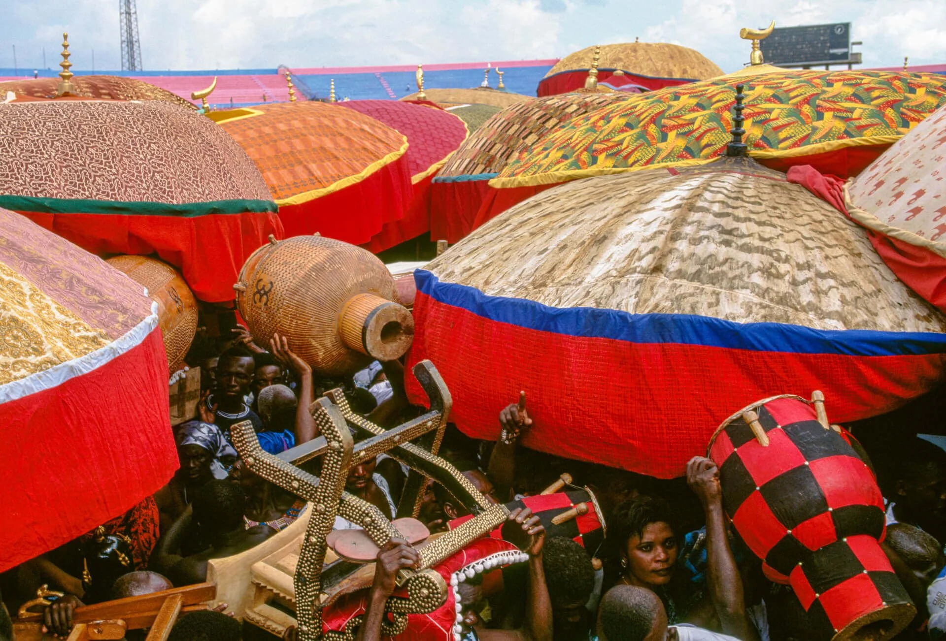 Ashanti umbrellas shielding paramount chiefs, Ghana. This image shows a gathering of Ashanti paramount chiefs arriving to honor the king, each sheltered beneath large ceremonial umbrellas crafted from richly patterned silk damask. These towering umbr