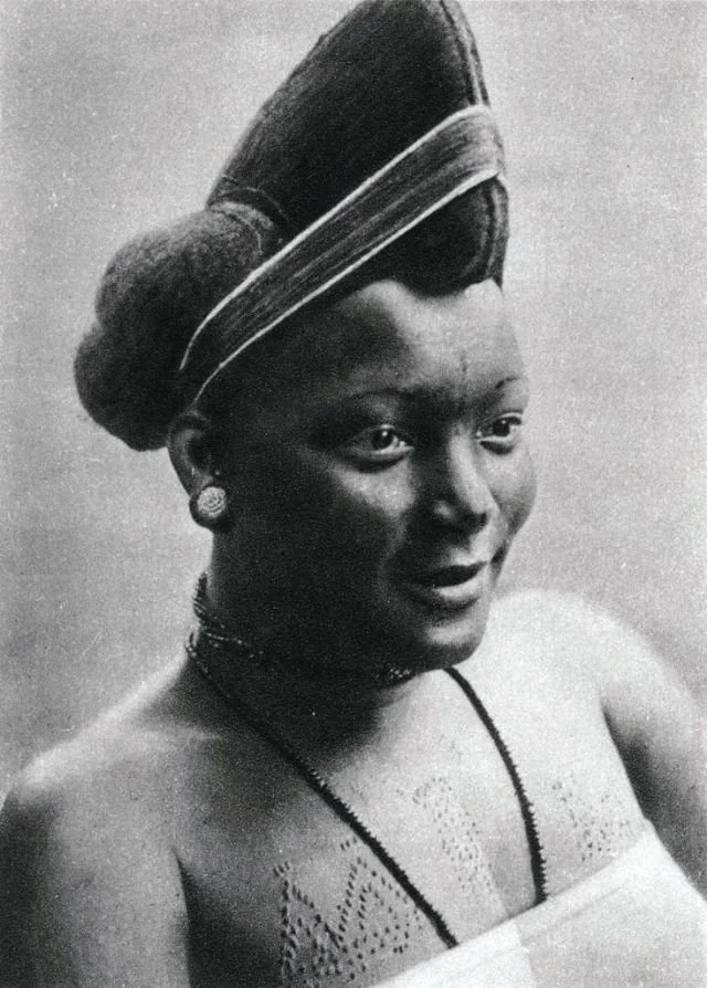 A structured, cone-shaped sculptural hairstyle with a voluminous rear roll, worn by a royal woman from Foumban, Cameroon circa 1911. Sourced from Vintag.es.