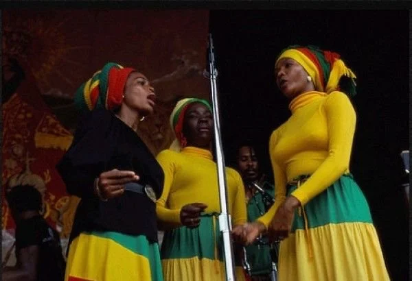 Caribbean women performing in Rastafarian-inspired attire, wearing red, yellow, and green headwraps and skirts.
