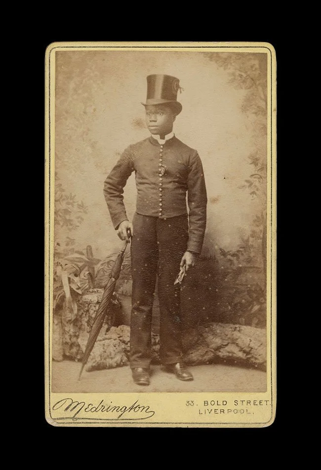 Full-length portrait of a Black boy wearing a top hat and holding an umbrella, posed before a painted landscape backdrop. Photographed by Medrington, Liverpool, England (Bold Street) , circa 1870s–1880s.